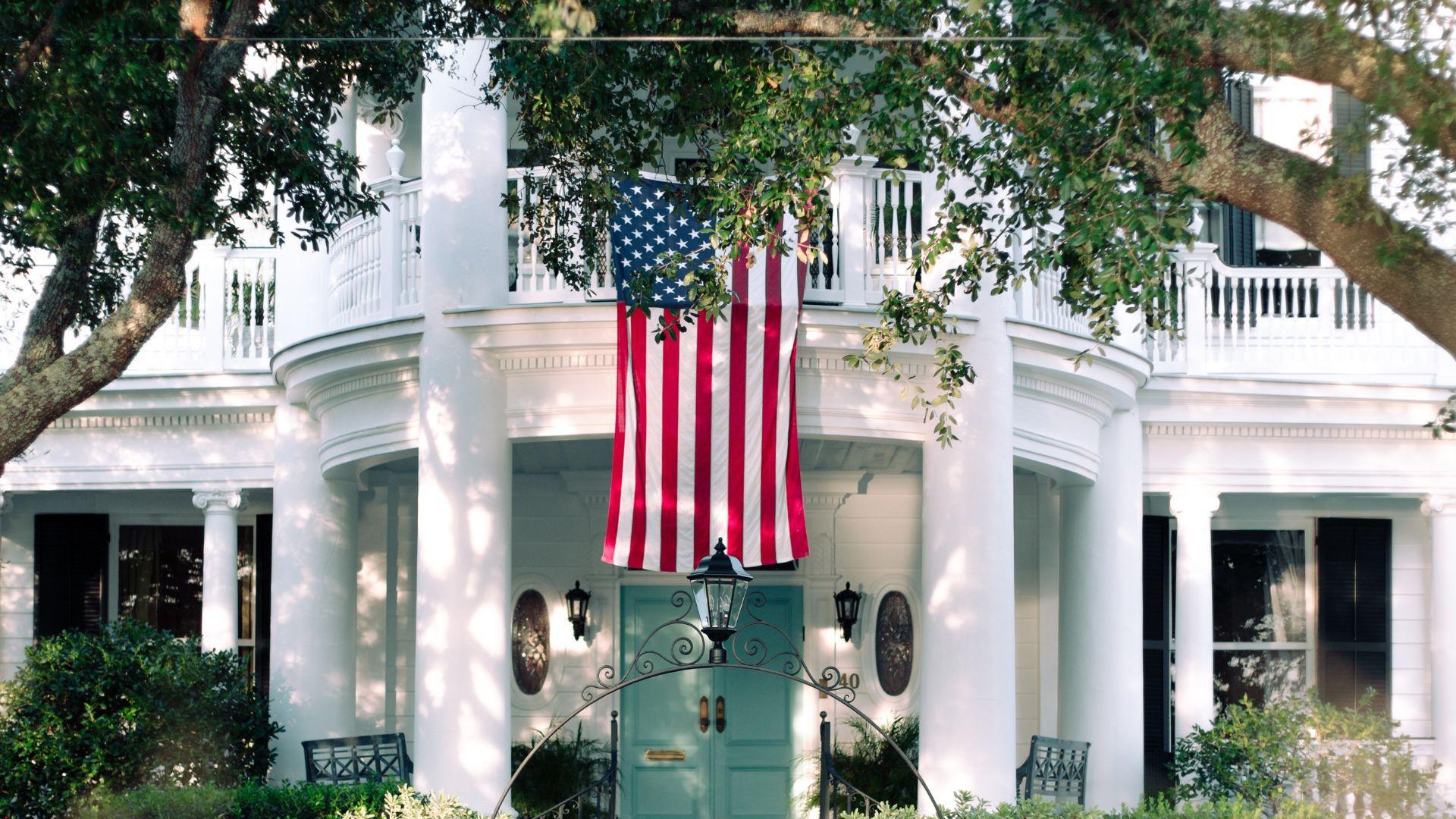 A two-story white house with columns, a blue front door, and a large American flag hanging in front
