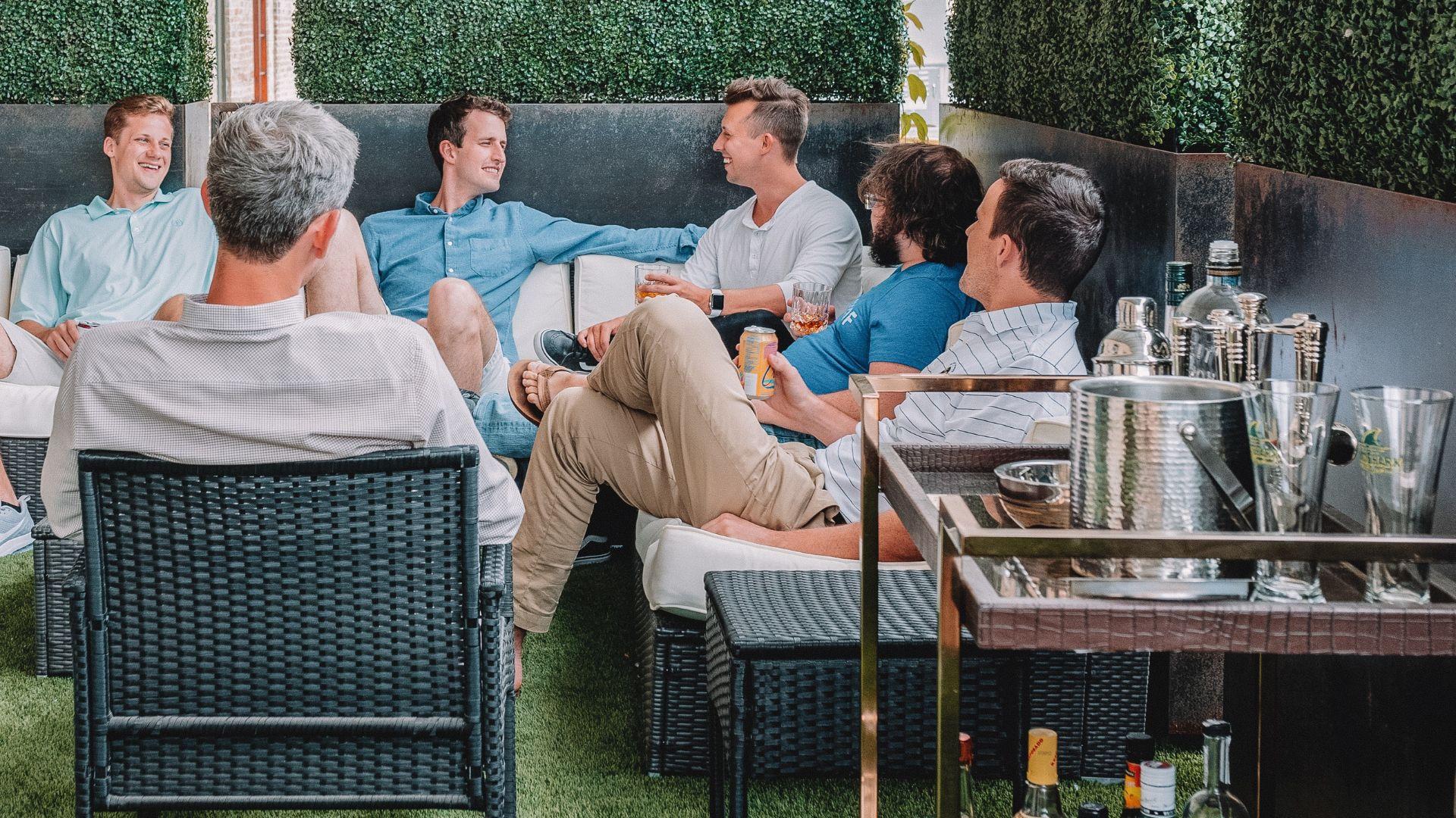 Group of men seated and chatting in an outdoor setting with plush sofas, surrounded by green hedges