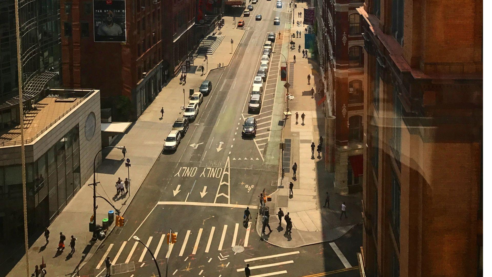 Aerial perspective of a city intersection with modern and brick buildings. Cars are parked alongside the road, and pedestrians cross the street