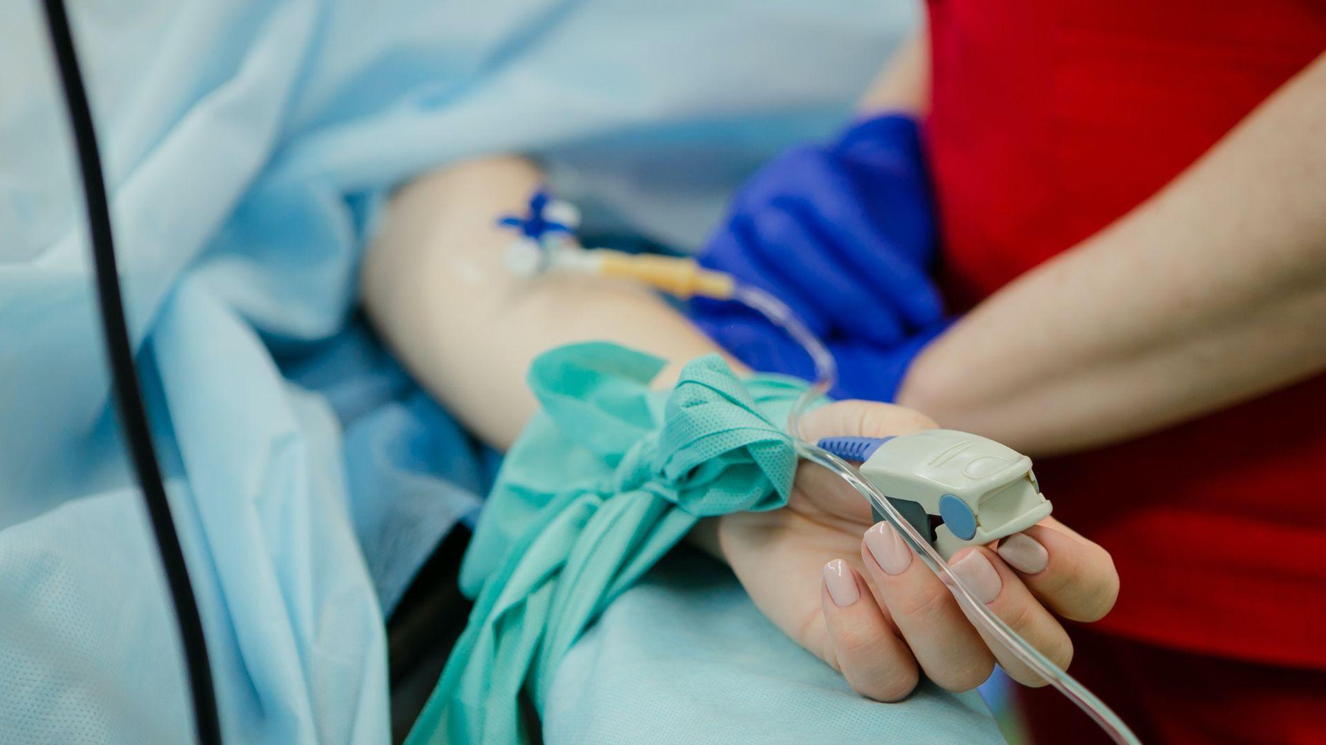 Close-up of a hand holding a medical device next to a patient's arm, which is covered with a blue sterile drape. An intravenous (IV) line is inserted into the patient's arm, and the background shows hints of medical equipment