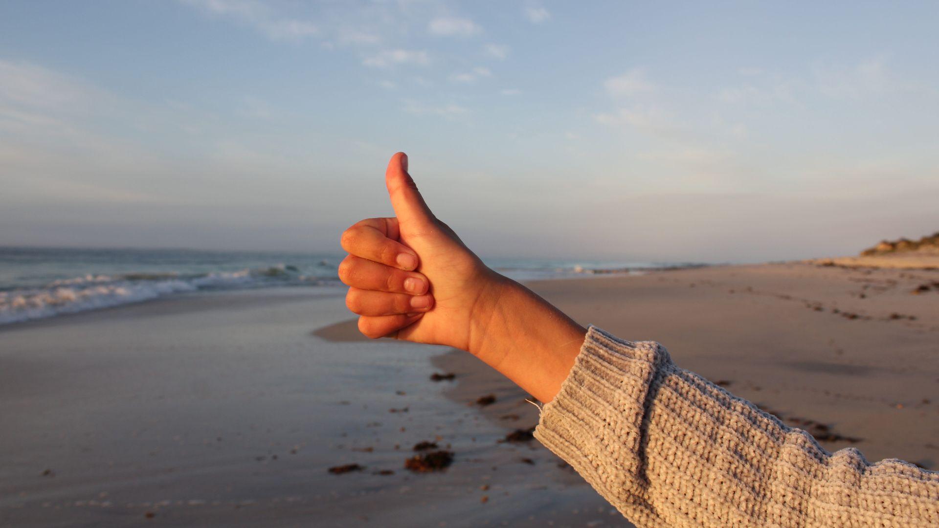 Close-up of a hand giving a thumbs-up against the backdrop of a sandy beach