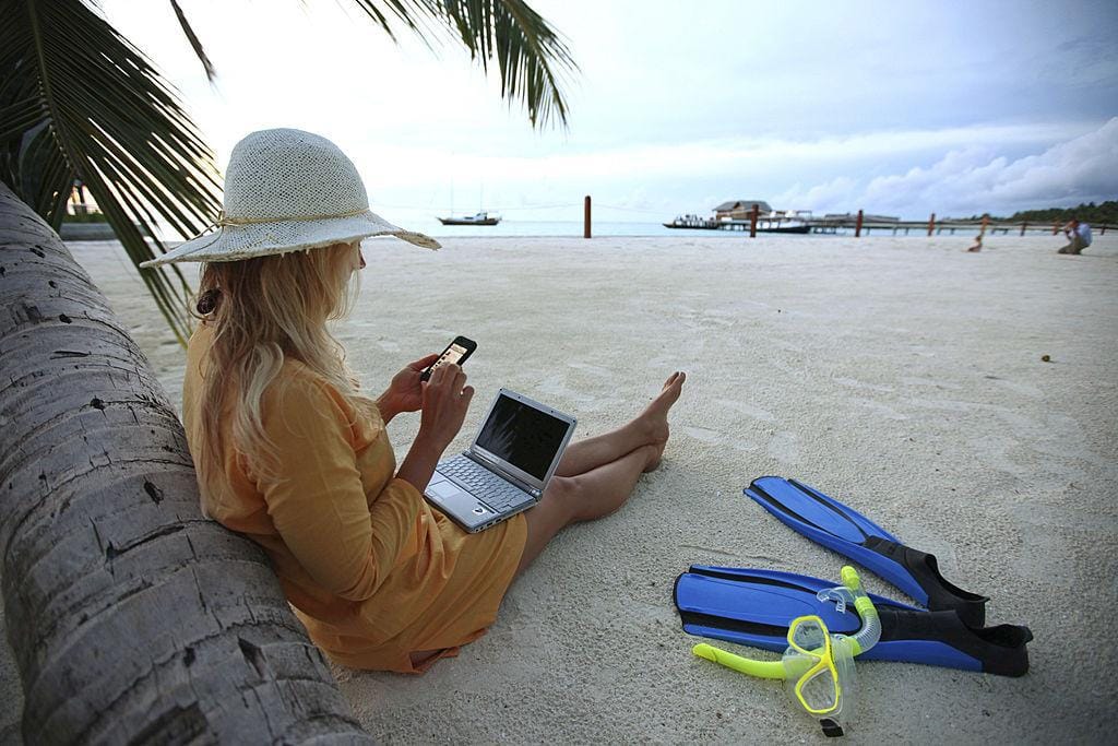 Woman sitting on the beach looking at her phone with her laptop on her lap